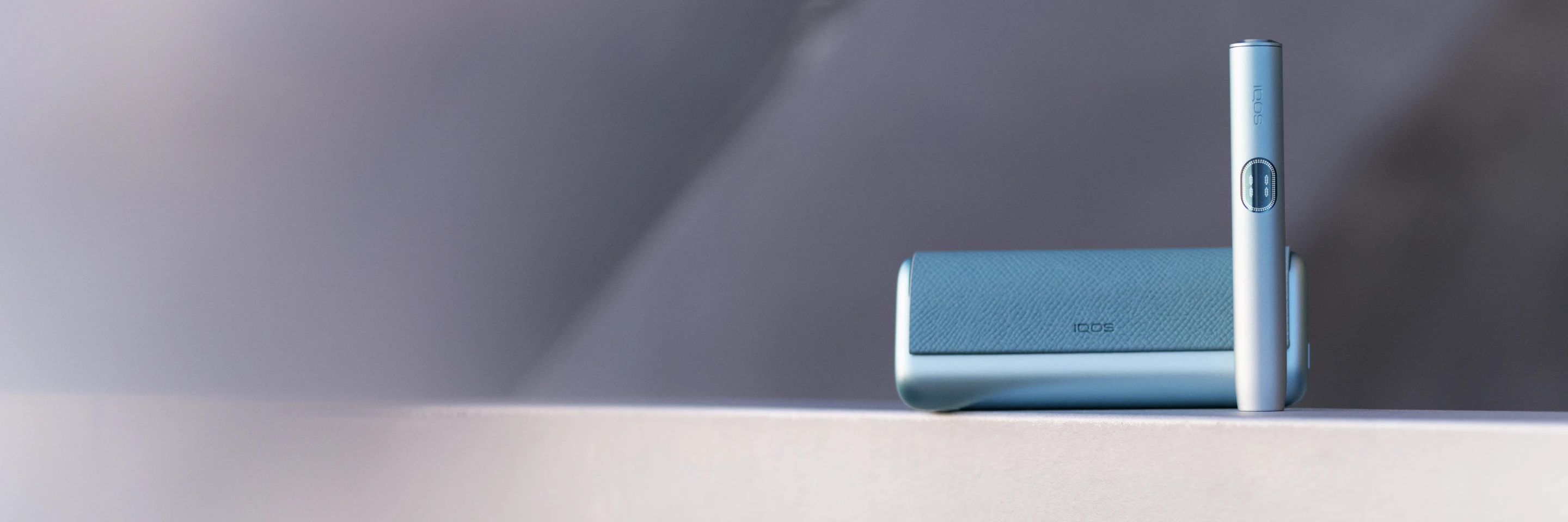 IQOS ILUMA i PRIME Breeze blue spalvos laikiklis ir kišeninis įkroviklis pilkame fone su užrašu „Įrenginio SPA paslaugos tavo IQOS“ ir mygtuku „Atrasti daugiau“.