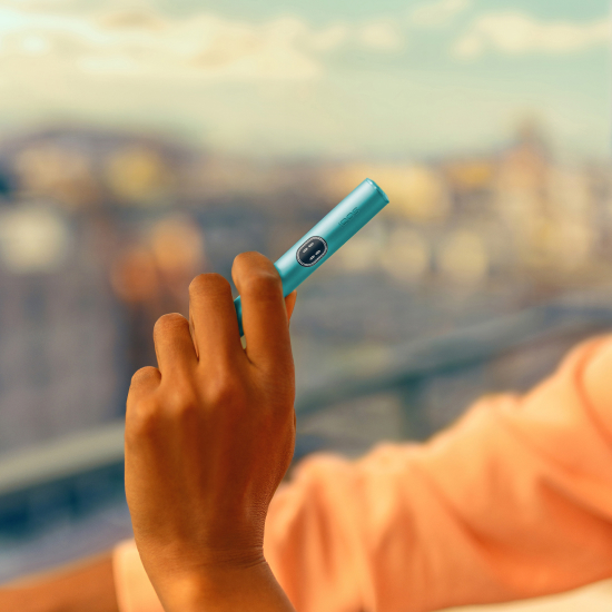 A hand holding an IQOS ILUMA i holder in Breeze blue color with a city view in the background
