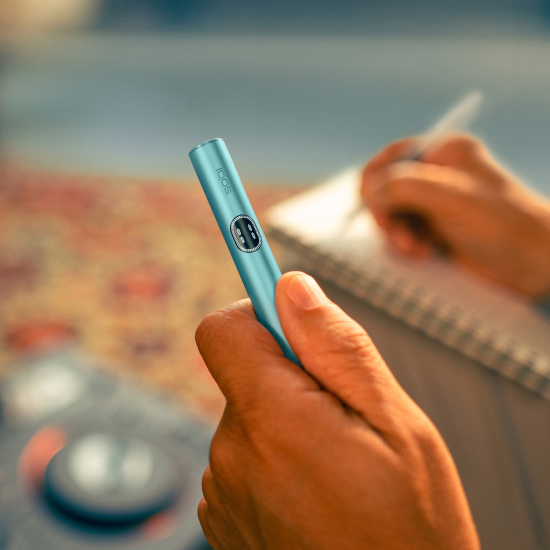 A hand holding an IQOS ILUMA i holder in Breeze Blue color, a notebook in the background.