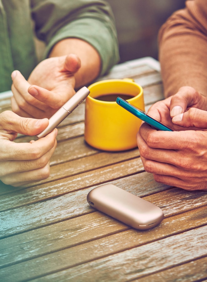 Two people talking at a table bar, one smokes IQOS and the other uses his phone